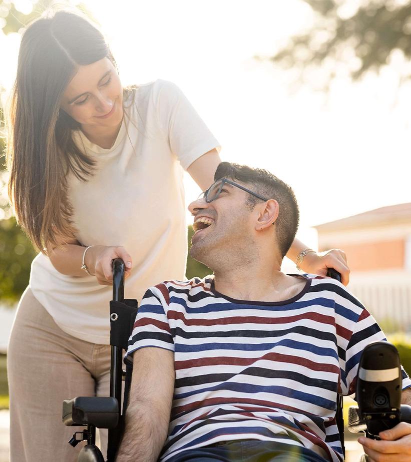 Caring woman pushes smiling man in wheelchair outdoors during sunny afternoon walk