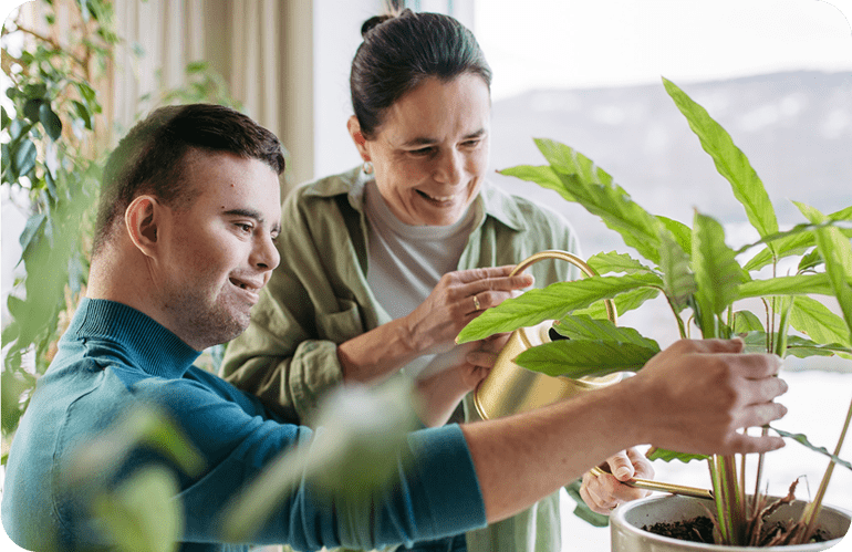Caregiver and young adult watering an indoor plant together.