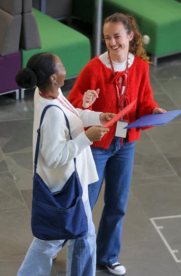 Two professionals wearing ID badges smile while discussing documents in a modern office lobby, holding red and blue folders during a conversation.