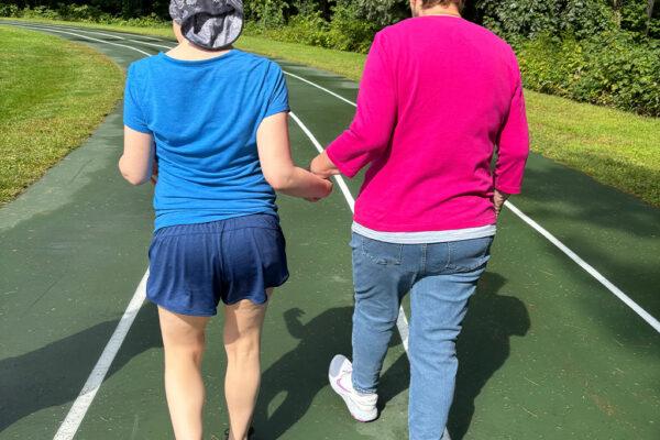 Two people walking hand in hand on an outdoor tennis court surrounded by trees and greenery.